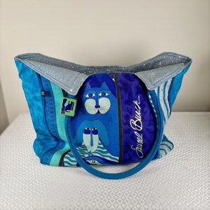 May include: A blue tote bag with a cat and dog design. The bag features a variety of blue shades, including a textured turquoise, and a deep blue velvet panel with the text "Faurel Burch". The bag has a blue handle and a light blue interior.