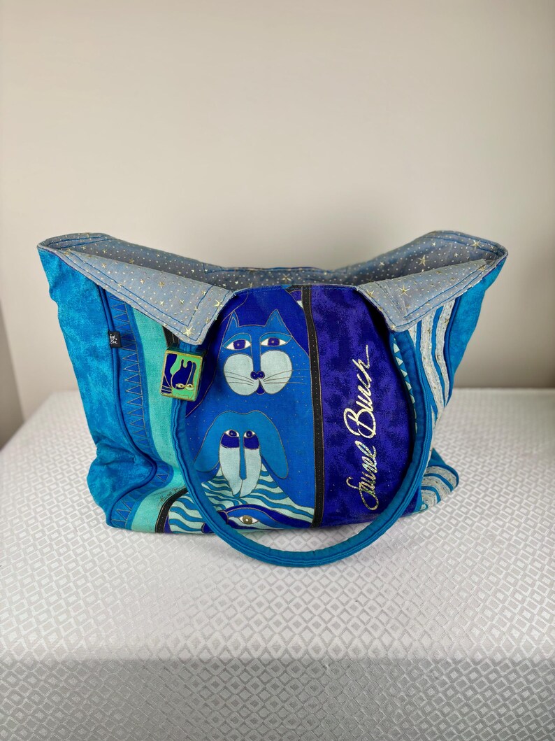 May include: A blue tote bag with a cat and dog design. The bag features a variety of blue shades, including a textured turquoise, and a deep blue velvet panel with the text "Faurel Burch". The bag has a blue handle and a light blue interior.
