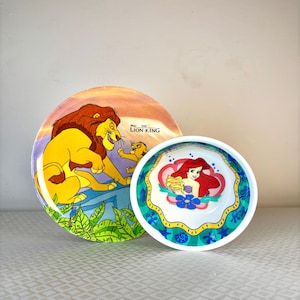 Op de afbeelding: Twee decoratieve borden met Disney-personages. E&eacute;n bord heeft een sc&egrave;ne uit "The Lion King" met Simba en Mufasa. Het andere bord heeft Ariel uit "The Little Mermaid". Beide borden zijn felgekleurd.