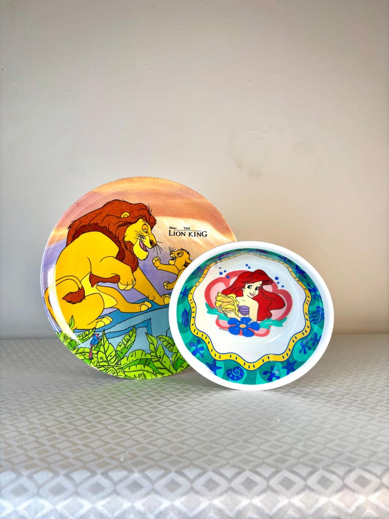 Op de afbeelding: Twee decoratieve borden met Disney-personages. E&eacute;n bord heeft een sc&egrave;ne uit "The Lion King" met Simba en Mufasa. Het andere bord heeft Ariel uit "The Little Mermaid". Beide borden zijn felgekleurd.