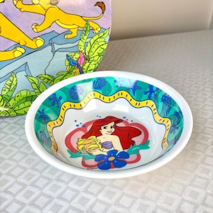 Op de afbeelding: Witte ovale kom met Ariel uit Disney's De Kleine Zeemeermin. De kom heeft een kleurrijke rand met een geel golvend ontwerp en blauwe en groene accenten. De kom staat voor een Lion King boek.