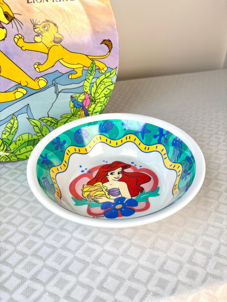 Op de afbeelding: Witte ovale kom met Ariel uit Disney's De Kleine Zeemeermin. De kom heeft een kleurrijke rand met een geel golvend ontwerp en blauwe en groene accenten. De kom staat voor een Lion King boek.