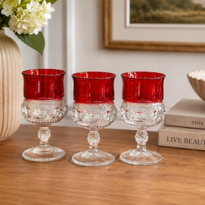Puede incluir: Tres copas de cristal transparente con la parte superior roja. Las copas tienen un diseño decorativo en la mitad inferior. Están sobre una superficie de madera. Un jarrón con flores blancas está en el fondo.