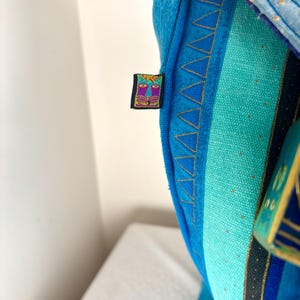 May include: Close-up of a blue textile with a colorful embroidered label. The textile features a variety of blue shades, with gold stitching and a geometric pattern. The label has a face design with purple and yellow accents.