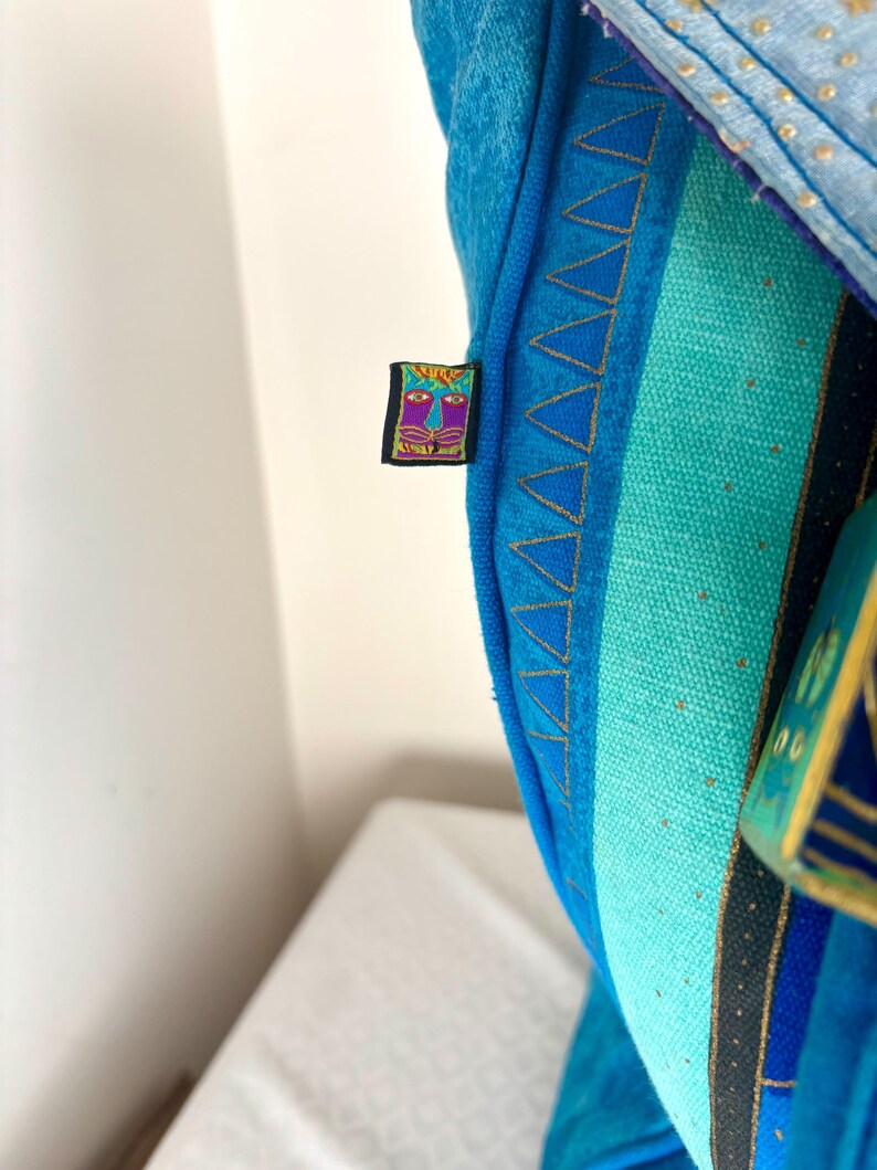 May include: Close-up of a blue textile with a colorful embroidered label. The textile features a variety of blue shades, with gold stitching and a geometric pattern. The label has a face design with purple and yellow accents.