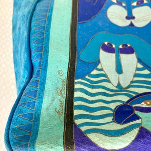 May include: A blue tote bag with a whimsical cat and dog design. The bag features a light blue background with a turquoise stripe and a black border. The artwork includes shades of blue, purple, and gold. The bag is made of a sturdy material.