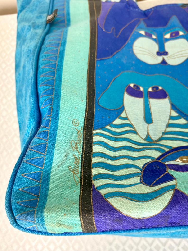 May include: A blue tote bag with a whimsical cat and dog design. The bag features a light blue background with a turquoise stripe and a black border. The artwork includes shades of blue, purple, and gold. The bag is made of a sturdy material.