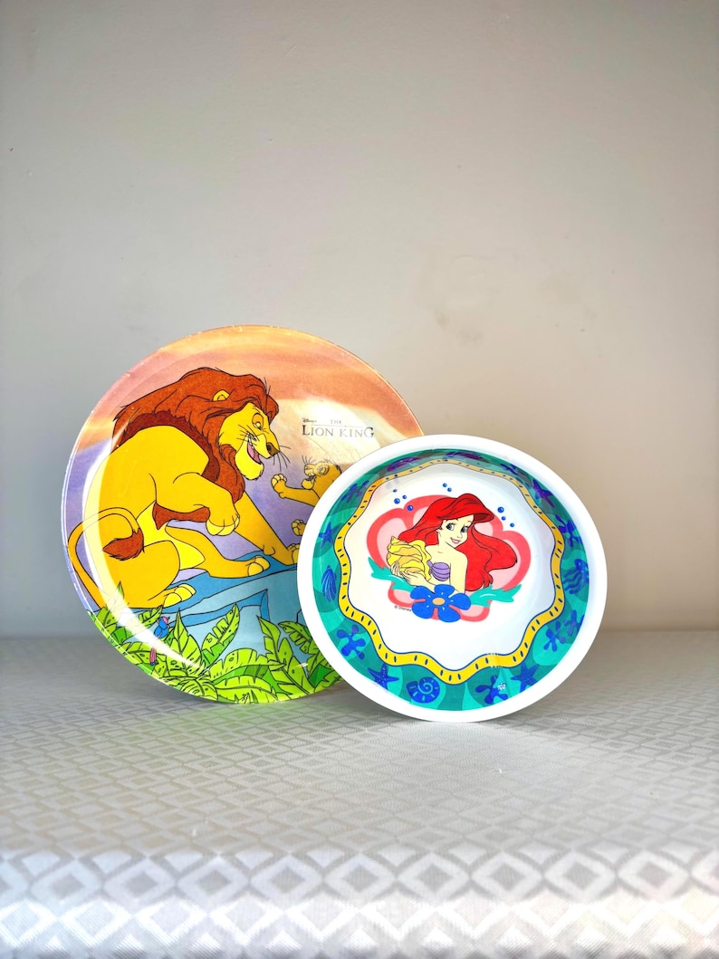 Op de afbeelding: Twee decoratieve borden met Disney-personages. E&eacute;n bord toont Simba uit The Lion King, met een gele leeuw en een zonsondergangachtergrond. Het andere bord toont Ariel uit The Little Mermaid, met een roodharige zeemeermin.