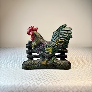May include: A cast iron cockerel figurine standing in front of a horizontal fence section. The cockerel features a bright red comb and wattle, with dark green, black, and yellow plumage. The base is textured to resemble grass, and the entire piece has a weathered, multi-coloured finish.