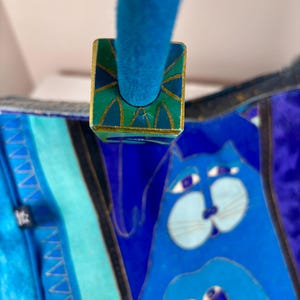 May include: A blue handbag with a whimsical cat design. The bag features a teal handle and a decorative square clasp with gold trim and a green and teal geometric pattern. The cat illustration is in shades of blue.