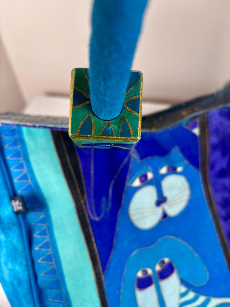 May include: A blue handbag with a whimsical cat design. The bag features a teal handle and a decorative square clasp with gold trim and a green and teal geometric pattern. The cat illustration is in shades of blue.