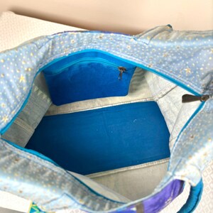 May include: Open view of a blue tote bag with a celestial pattern of gold stars on a light blue background. The interior features a vibrant blue pocket with a zipper and a solid blue base. The handles are a bright turquoise color.