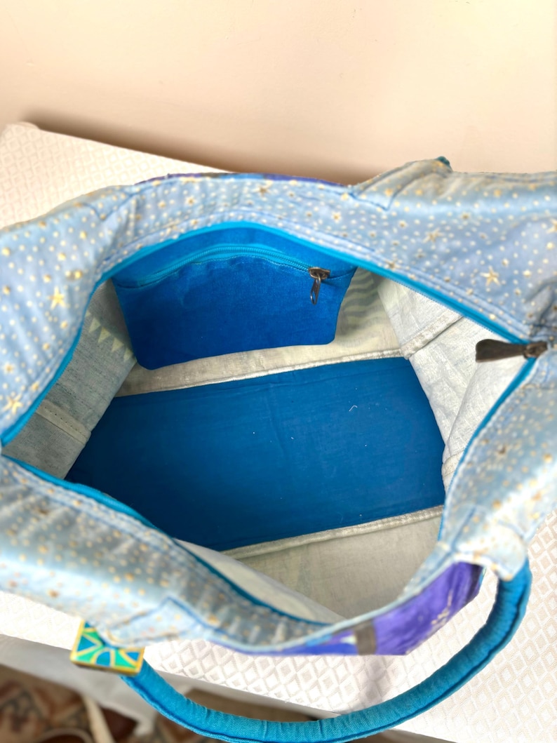 May include: Open view of a blue tote bag with a celestial pattern of gold stars on a light blue background. The interior features a vibrant blue pocket with a zipper and a solid blue base. The handles are a bright turquoise color.
