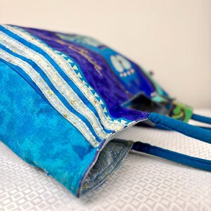 May include: A blue tote bag with a vibrant, abstract design. The bag features a mix of blue hues, with white and gold accents. The handles are a solid blue, and the interior lining is a lighter blue with gold flecks. The bag is made of fabric.