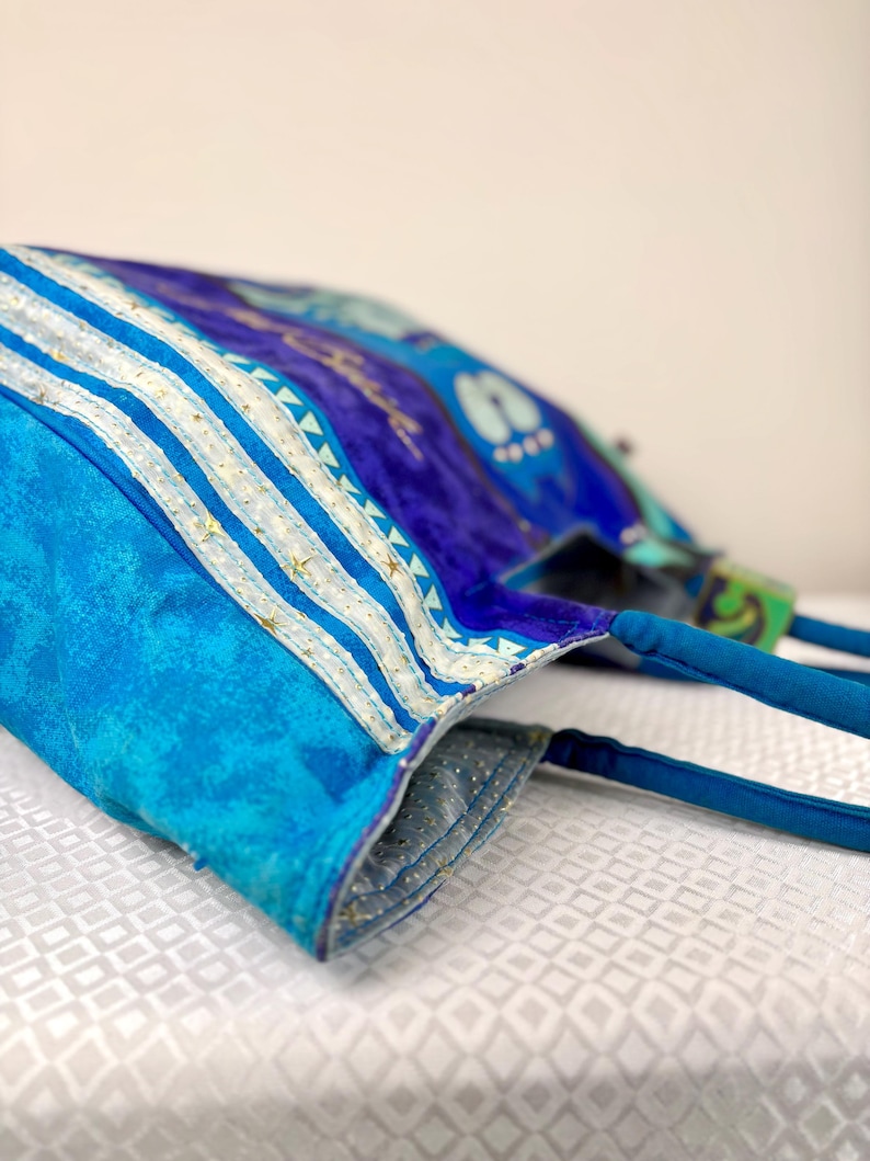 May include: A blue tote bag with a vibrant, abstract design. The bag features a mix of blue hues, with white and gold accents. The handles are a solid blue, and the interior lining is a lighter blue with gold flecks. The bag is made of fabric.
