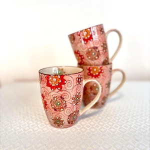 May include: Three stacked ceramic mugs with floral patterns in shades of pink, red, orange, and green. The mugs have cream-colored handles and are set against a white background.