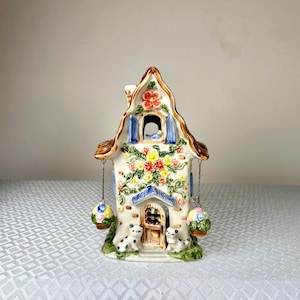 May include: A ceramic house-shaped candle holder with a brown roof, white walls, and colorful floral and vine decorations. The house has a door, window, and chimney. Two small hanging baskets and two small cat figurines are also visible.