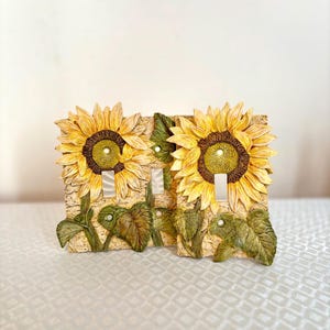 Puede incluir: Cubierta decorativa para interruptor de luz con dos girasoles de pétalos amarillos y centros marrones. La cubierta también incluye hojas verdes y está montada sobre un fondo beige texturizado. Los interruptores son blancos.