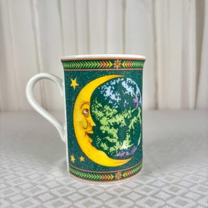 Puede incluir: Una taza de cerámica blanca con fondo verde oscuro, decorada con una luna creciente amarilla con rostro y un gráfico de la Tierra en tonos verdes y azules. El diseño incluye pequeñas estrellas amarillas y un borde con patrones geométricos.