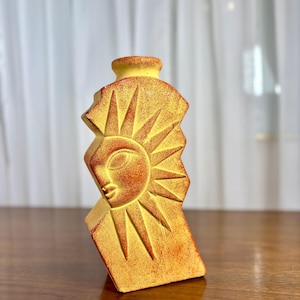 Könnte beinhalten: Eine kleine, aufrechte, gelb-orange texturierte Vase mit einer flachen Silhouette und gezackten Kanten. Die Vorderseite zeigt ein geschnitztes, stilisiertes Sonnenmotiv mit einem menschenähnlichen Gesichtsprofil und Strahlen.