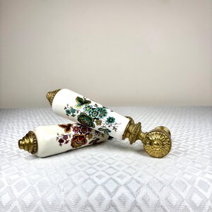May include: Two decorative porcelain drawer pulls with floral designs. The pulls have a white base with colorful flowers and gold-colored metal accents. One pull features green and blue flowers, while the other has red and yellow flowers.