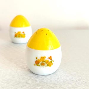 May include: Two egg-shaped salt and pepper shakers. The top half of each shaker is bright yellow, and the bottom half is white. A floral design with yellow, orange, and white flowers and a butterfly decorates the white section.