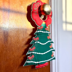 以下が含まれることがあります： 緑色の布製クリスマスツリーに赤いリボンと白いガーランドが付いたクリスマスドアハンガー。ツリーは「HAPPY」の文字が刺繍された赤いリースの下に吊り下げられています。 金色のドアノブに取り付けられています。