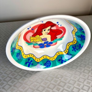 Op de afbeelding: Witte ovale keramische kom met een cartoon illustratie van Ariel uit De Kleine Zeemeermin. Het ontwerp bevat Ariel met rood haar, een paarse schelp top en een gele handdoek. De rand heeft een groen en gele oceaan-thema rand.