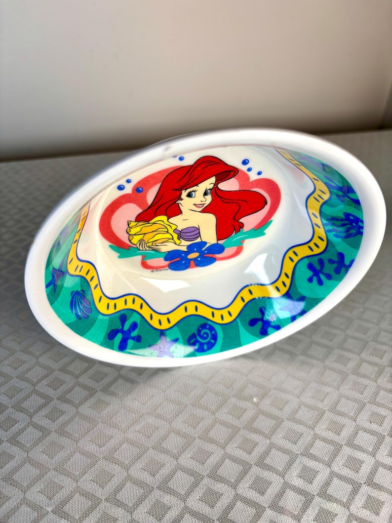 Op de afbeelding: Witte ovale keramische kom met een cartoon illustratie van Ariel uit De Kleine Zeemeermin. Het ontwerp bevat Ariel met rood haar, een paarse schelp top en een gele handdoek. De rand heeft een groen en gele oceaan-thema rand.
