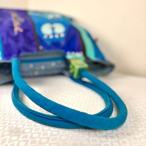 May include: A blue handbag with a soft, velvety texture. The bag features a vibrant design with shades of blue and green. The handles are a bright turquoise color, and the clasp is a light green. The bag is sitting on a white surface.