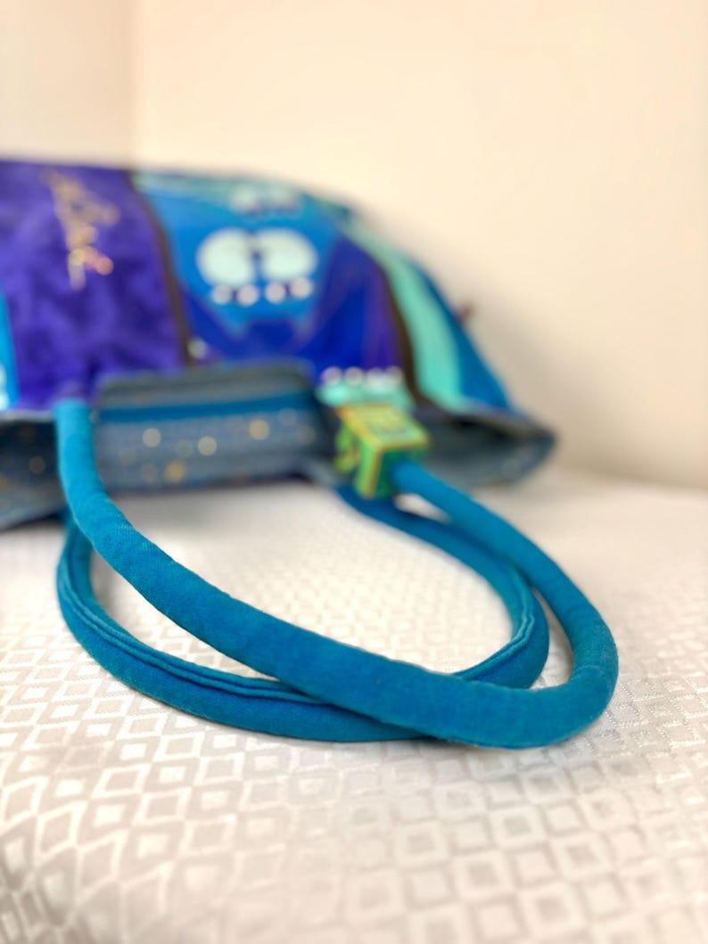 May include: A blue handbag with a soft, velvety texture. The bag features a vibrant design with shades of blue and green. The handles are a bright turquoise color, and the clasp is a light green. The bag is sitting on a white surface.