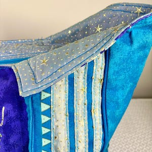 May include: Close-up of a blue and purple fabric bag with a celestial theme. The bag features gold star accents, vertical stripes, and a light blue lining. The design incorporates various shades of blue and purple.