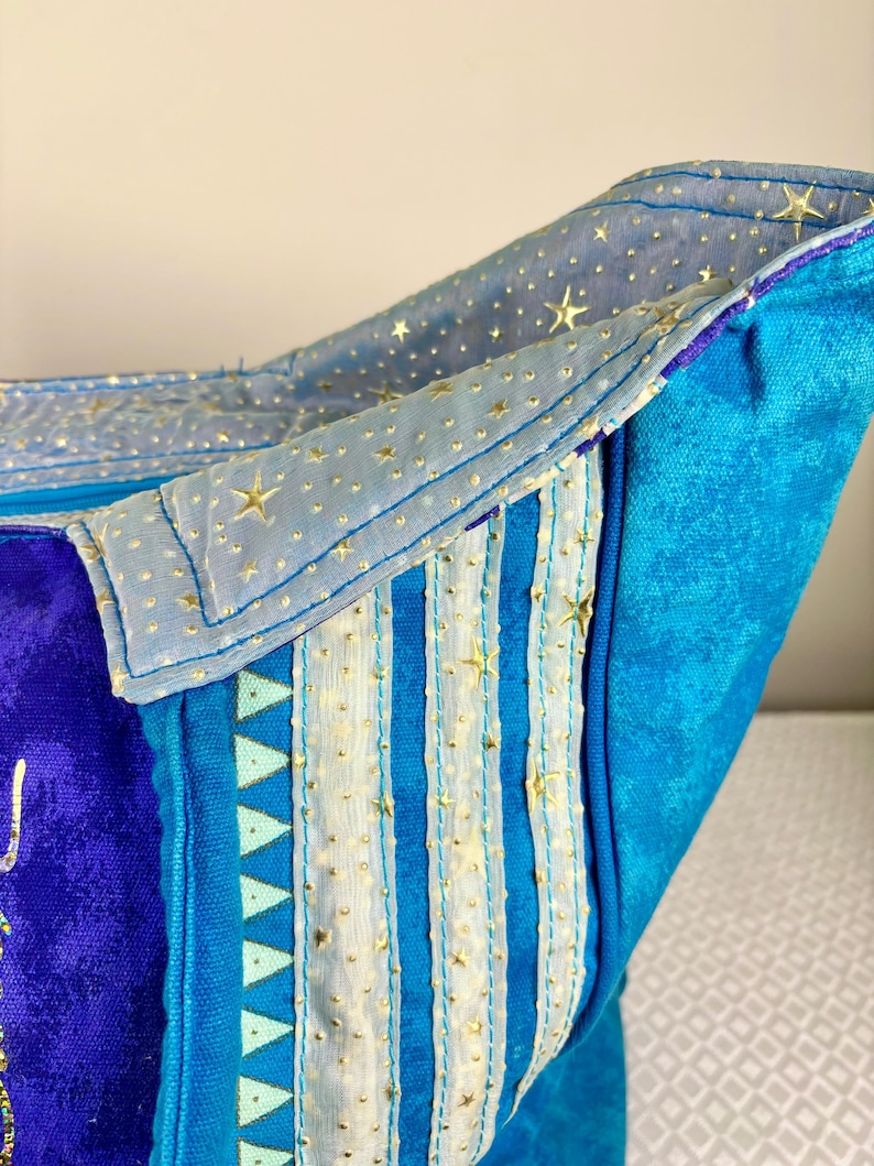 May include: Close-up of a blue and purple fabric bag with a celestial theme. The bag features gold star accents, vertical stripes, and a light blue lining. The design incorporates various shades of blue and purple.