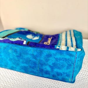 May include: A blue tote bag with a textured, suede-like exterior. The bag features a vibrant pattern in shades of blue, teal, and white. The word "Beach" is visible in cursive script. The bag has a rectangular shape.