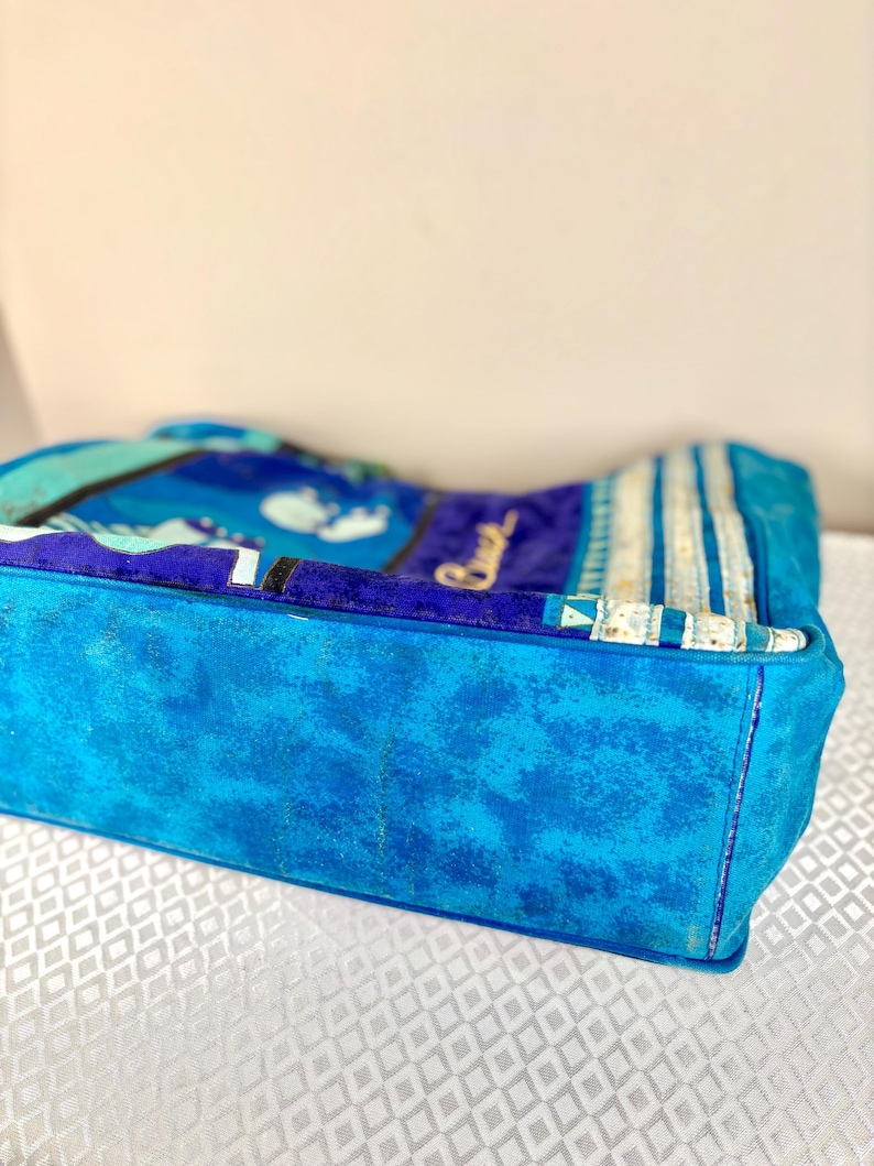 May include: A blue tote bag with a textured, suede-like exterior. The bag features a vibrant pattern in shades of blue, teal, and white. The word "Beach" is visible in cursive script. The bag has a rectangular shape.