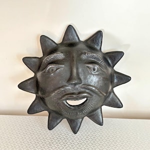 Könnte beinhalten: Ein dunkelgrauer, sonnenförmiger Wandbehang mit einem lächelnden Gesicht und spitzen Strahlen. Die Sonne hat einen Schnurrbart und Augenbrauen, und der Mund ist offen. Der Wandbehang besteht aus einem strukturierten Material.