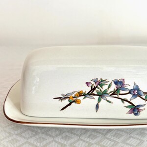 May include: A white ceramic butter dish with a lid, featuring a floral design in shades of blue, purple, yellow, and green. The dish has a brown trim around the edges. The butter dish is rectangular in shape.