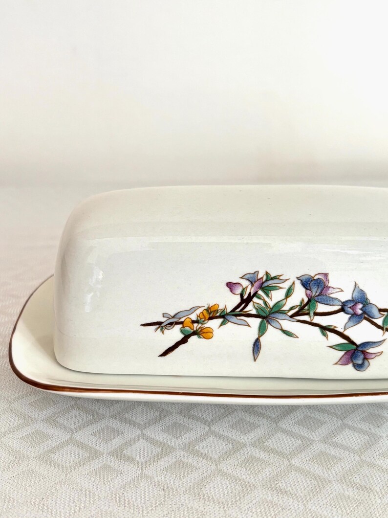 May include: A white ceramic butter dish with a lid, featuring a floral design in shades of blue, purple, yellow, and green. The dish has a brown trim around the edges. The butter dish is rectangular in shape.