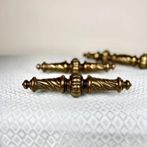 Może przedstawiać: Dwa mosiężne uchwyty do szuflad w stylu antycznym z centralnym żebrowanym elementem, otoczonym skręconymi, spiralnymi trzonami, które zwężają się ku zaokrąglonym zakończeniom. Metal ma wyblakłe, ciemnozłote wykończenie z widoczną teksturą.
