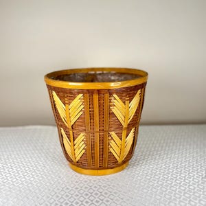 Puede incluir: Cesta cilíndrica de bambú tejido. La cesta es marrón y amarilla, con un patrón geométrico repetitivo. La base y el borde son amarillos, y el interior está forrado. Un artículo de almacenamiento decorativo.