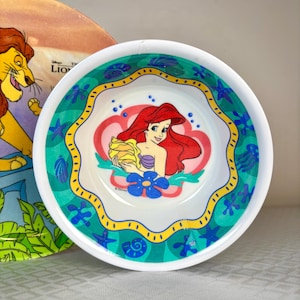 Op de afbeelding: Een witte kom met een kleurrijke illustratie van Ariel uit Disney's De Kleine Zeemeermin. De kom heeft een turquoise, gele en blauwe rand met schelp- en bloemmotieven. Een Lion King-bord is gedeeltelijk zichtbaar op de achtergrond.