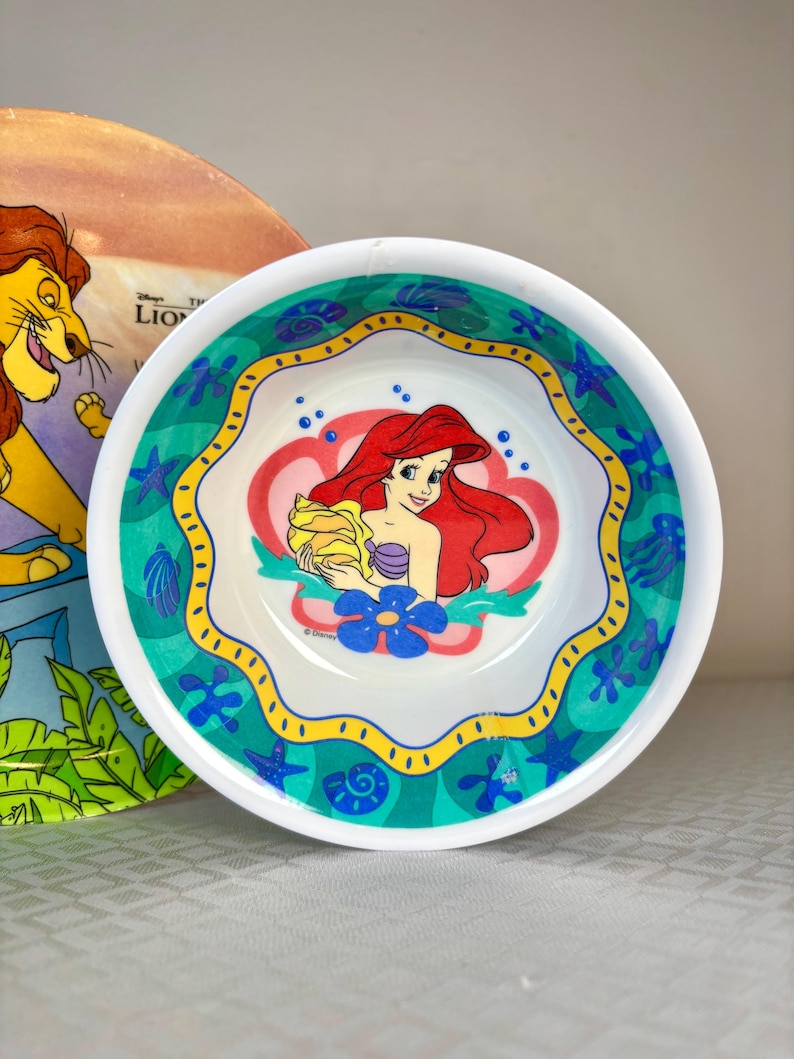 Op de afbeelding: Een witte kom met een kleurrijke illustratie van Ariel uit Disney's De Kleine Zeemeermin. De kom heeft een turquoise, gele en blauwe rand met schelp- en bloemmotieven. Een Lion King-bord is gedeeltelijk zichtbaar op de achtergrond.