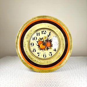 May include: A round vintage wall clock with a gold, orange, and brown frame. The clock face has a fruit design and black numerals. This decorative clock is a vintage item.