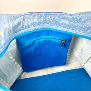 May include: A blue tote bag interior with a zippered pocket. The bag's lining features a light blue fabric with gold stars. The interior pocket is a solid blue, and the zipper is silver.