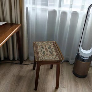 May include: A small wooden stool with a floral patterned cushion top. The stool is in a room with white curtains and a brown wooden table.