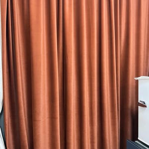 Könnte beinhalten: Nahaufnahme von rostfarbenen Samtvorhängen. Die Vorhänge hängen in vertikalen Falten und erzeugen ein strukturiertes Aussehen. Die Farbe ist ein warmer, erdiger Ton, und der Stoff wirkt weich und luxuriös. Die Vorhänge werden wahrscheinlich für die Inneneinrichtung verwendet.