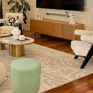 May include: A light green patterned ottoman with a geometric design. The ottoman is in a living room setting with a rug, a coffee table, and a chair.