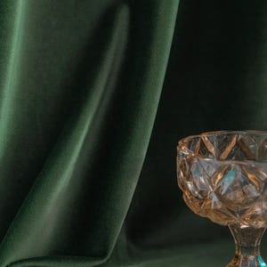 May include: A clear glass bowl with a faceted design sits on a dark green velvet surface. The bowl has a wide, shallow base and a tall, slender stem.