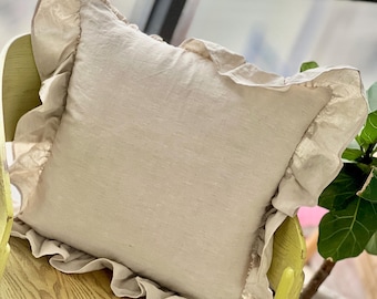 Puede incluir: Un cojín cuadrado de lino beige con un borde con volantes. El cojín está colocado sobre una silla de madera verde claro. El color neutro y la tela texturizada crean una estética suave y acogedora. El ribete con volantes añade un toque de elegancia.