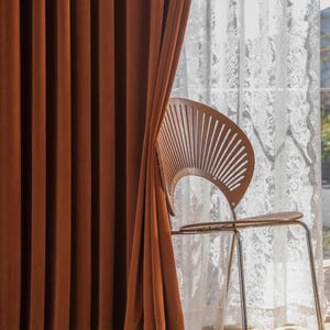May include: Rust-colored velvet curtains hang next to a white lace curtain. A wooden chair with a fan-shaped back and metal legs sits in front of the window. The curtains are drawn, partially obscuring the chair.